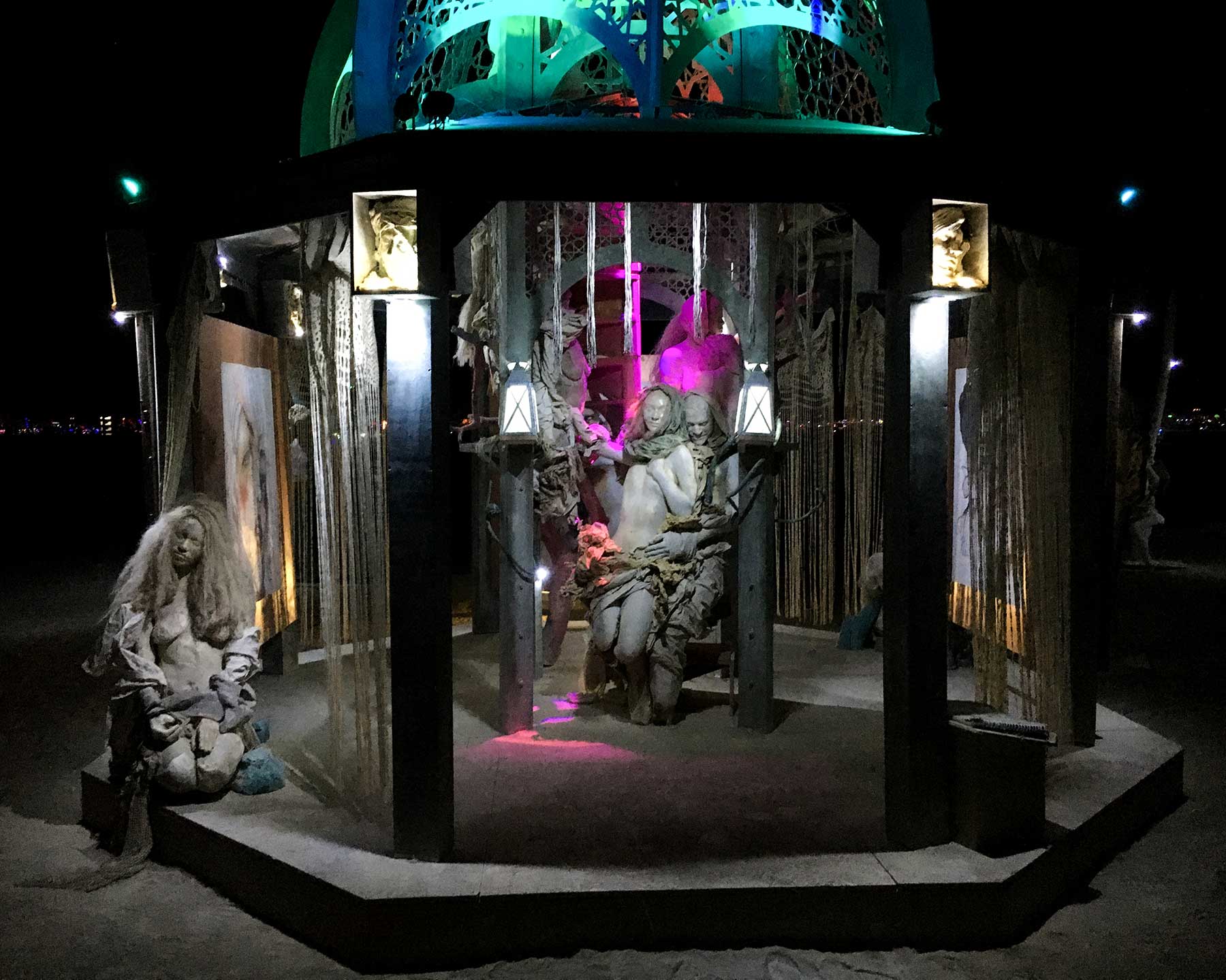 Night view into the structure: an embracing pair of cast figures lit in magenta between two lanterns, with a seated figure at left