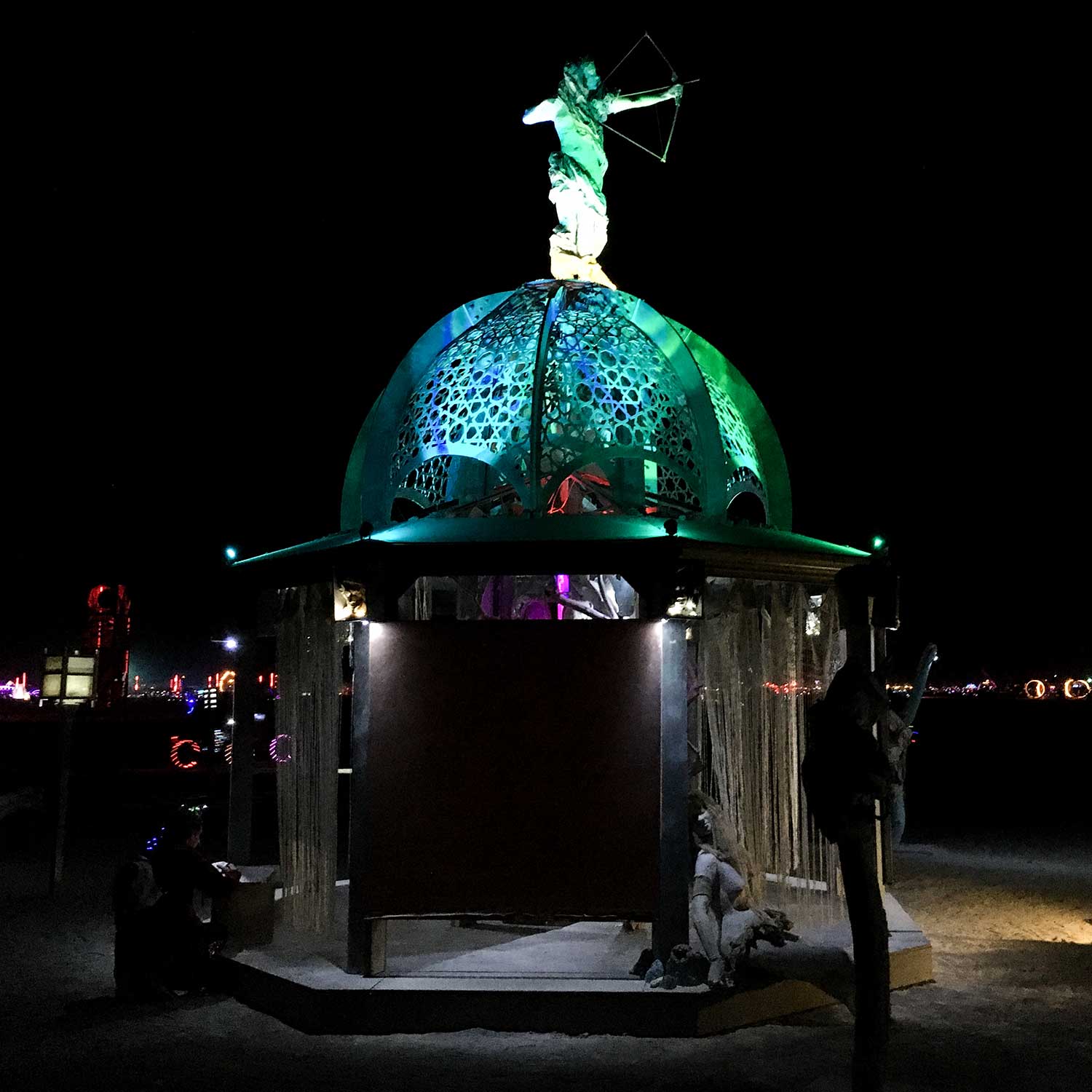 The structure at night from the side, its dome glowing green beneath the archer figure, with visitors in silhouette