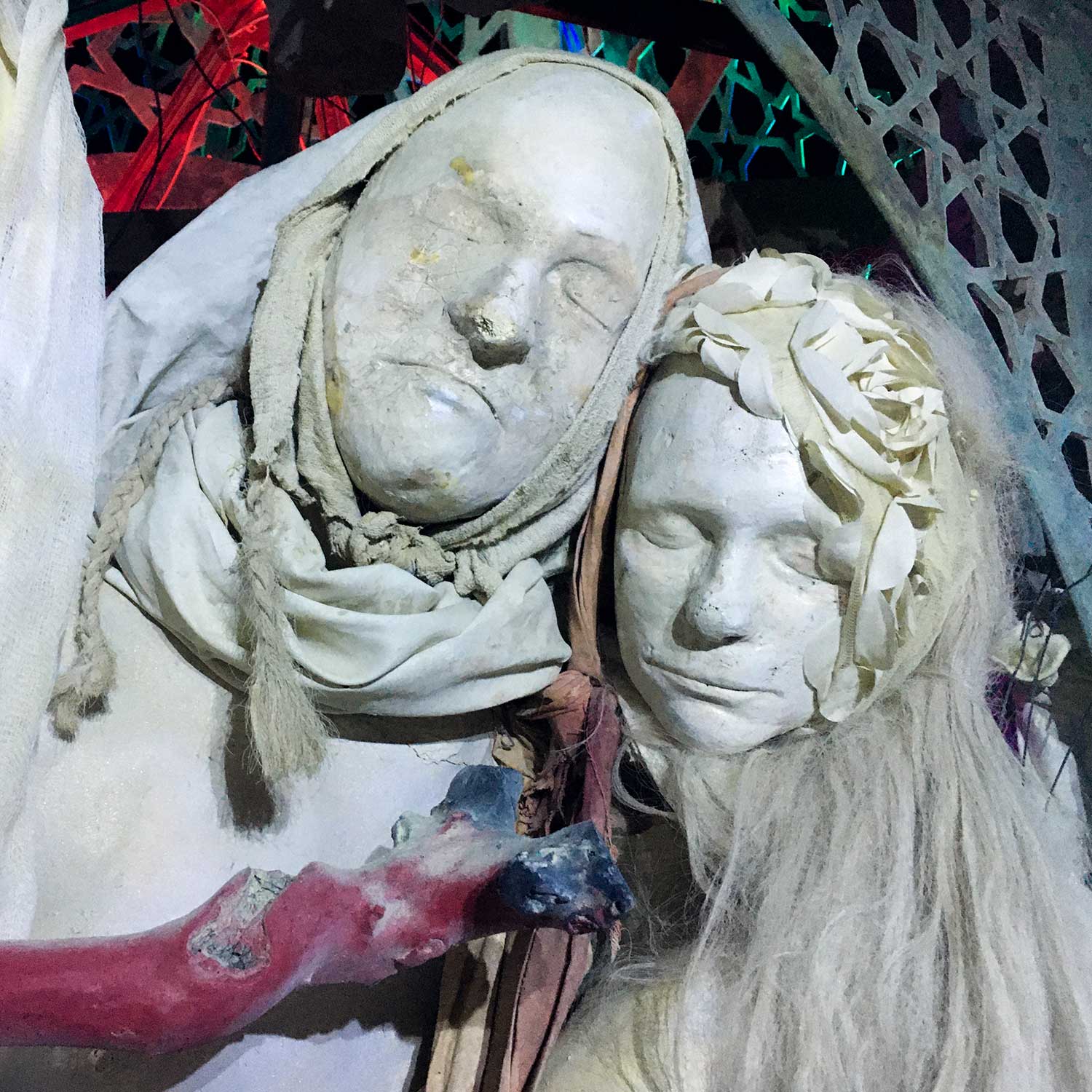 Close-up of two pale cast faces with closed eyes, one hooded, the other crowned with white flowers