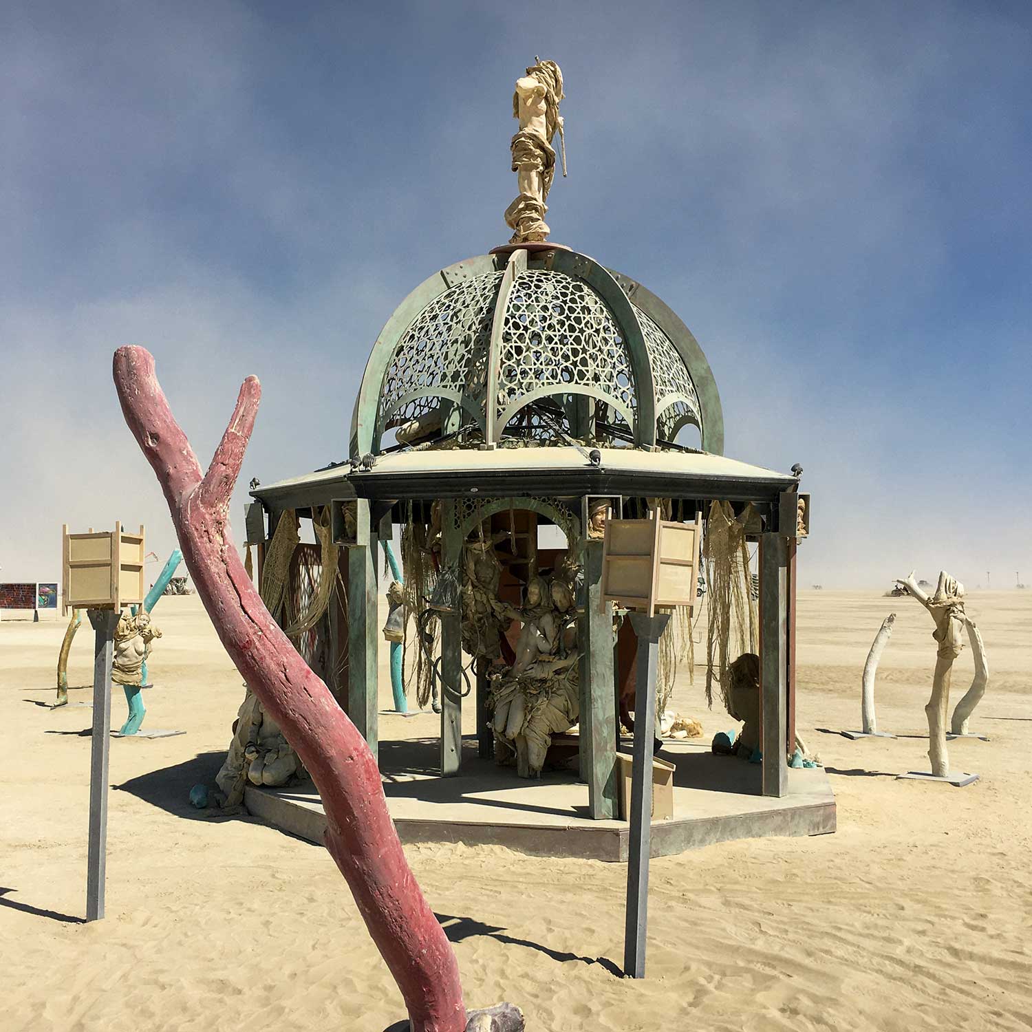 The structure by day with a red tree branch in the foreground, lantern posts and cast figures across the playa