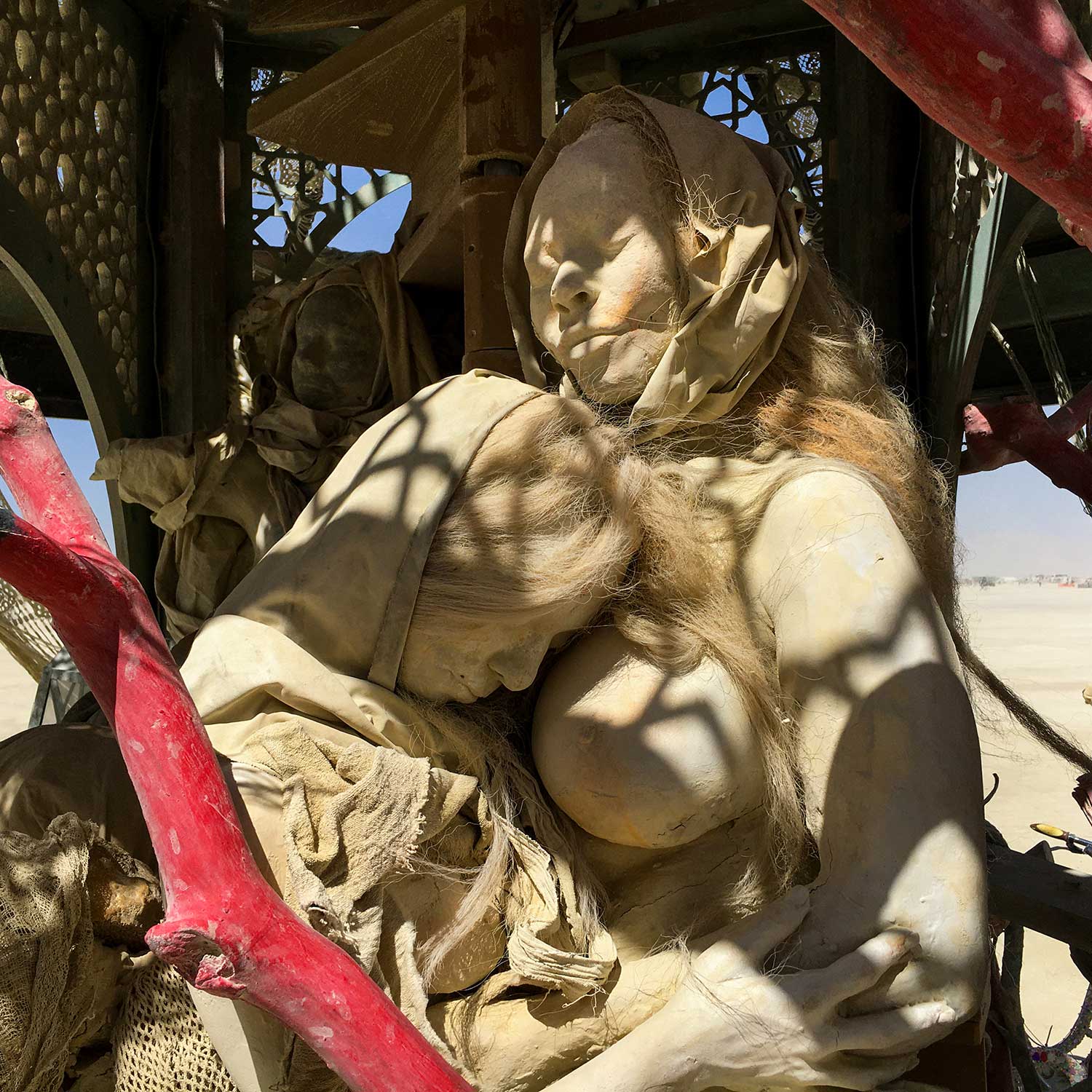 Close-up of a hooded cast mother cradling a long-haired figure, dappled by the shadow of the lattice dome