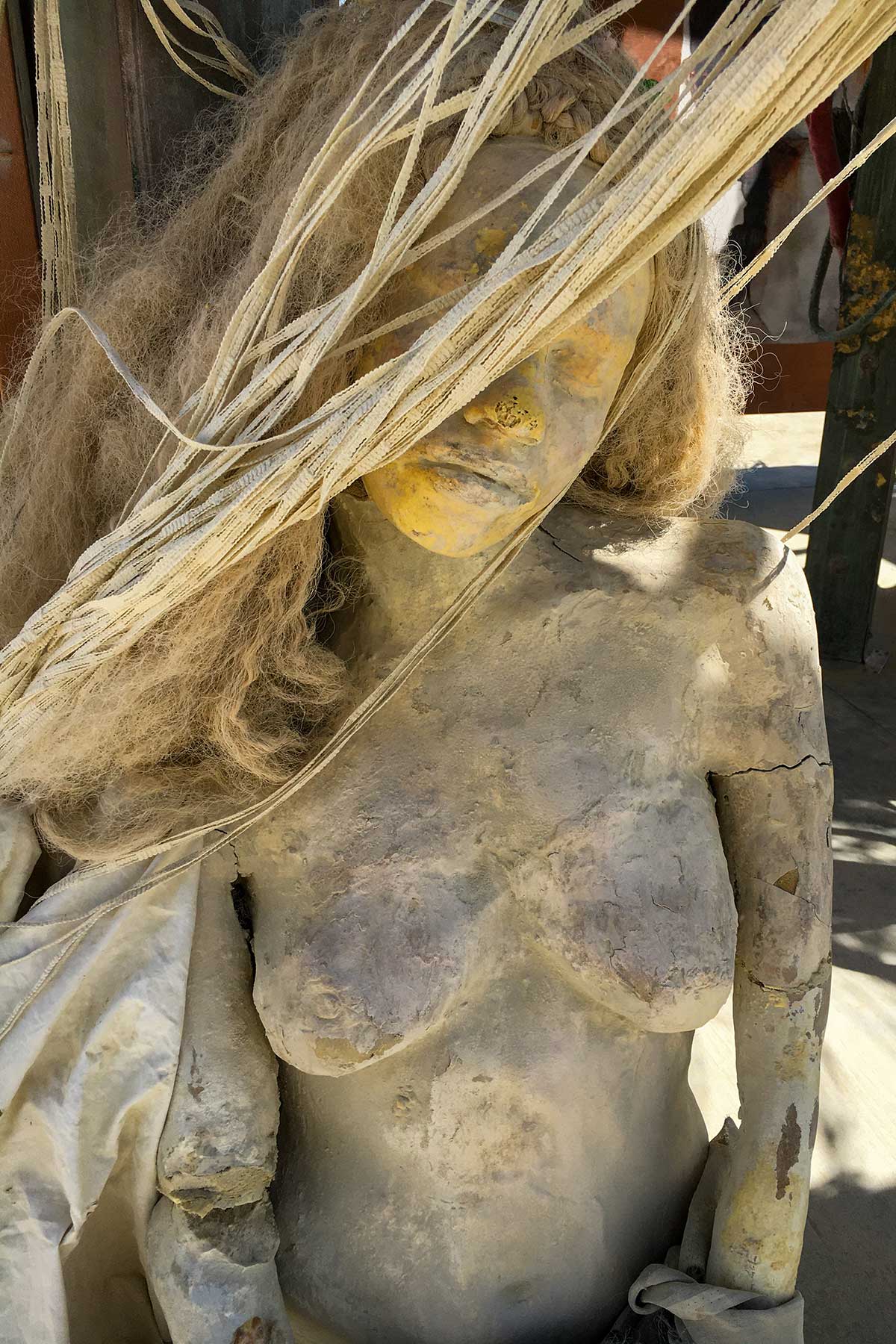 Close-up of a cracked, dust-covered cast figure with long hair, her face half hidden by hanging strands of cord