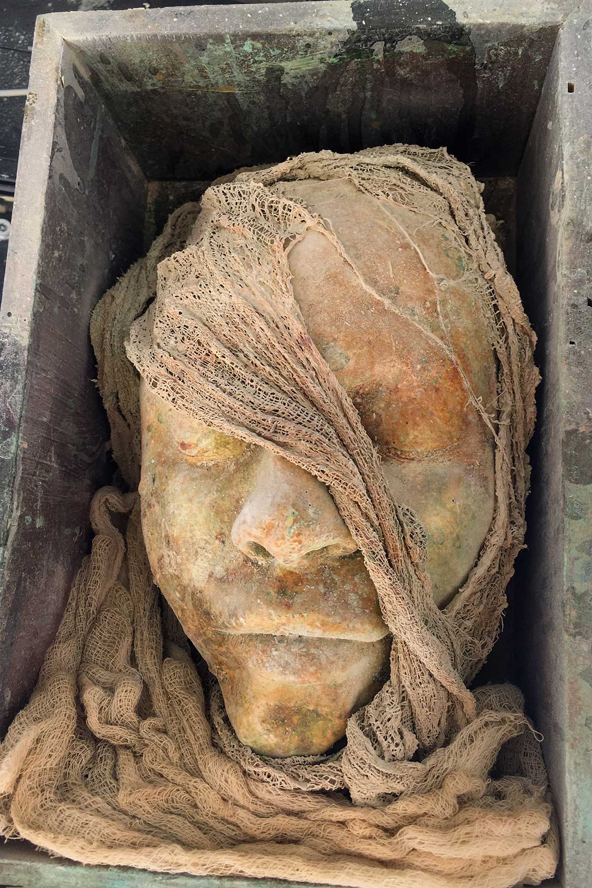 A cast face wrapped in gauze, resting in a weathered metal box
