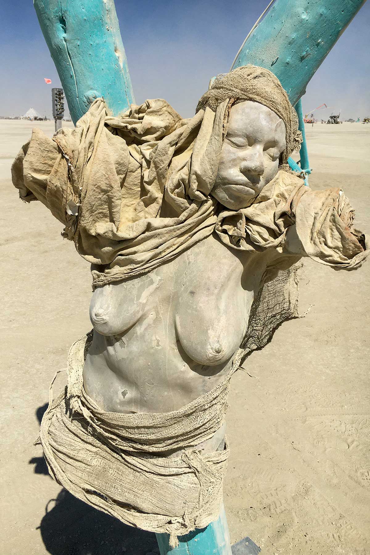 A cast figure with closed eyes, wrapped in sackcloth and fixed between the branches of a turquoise tree