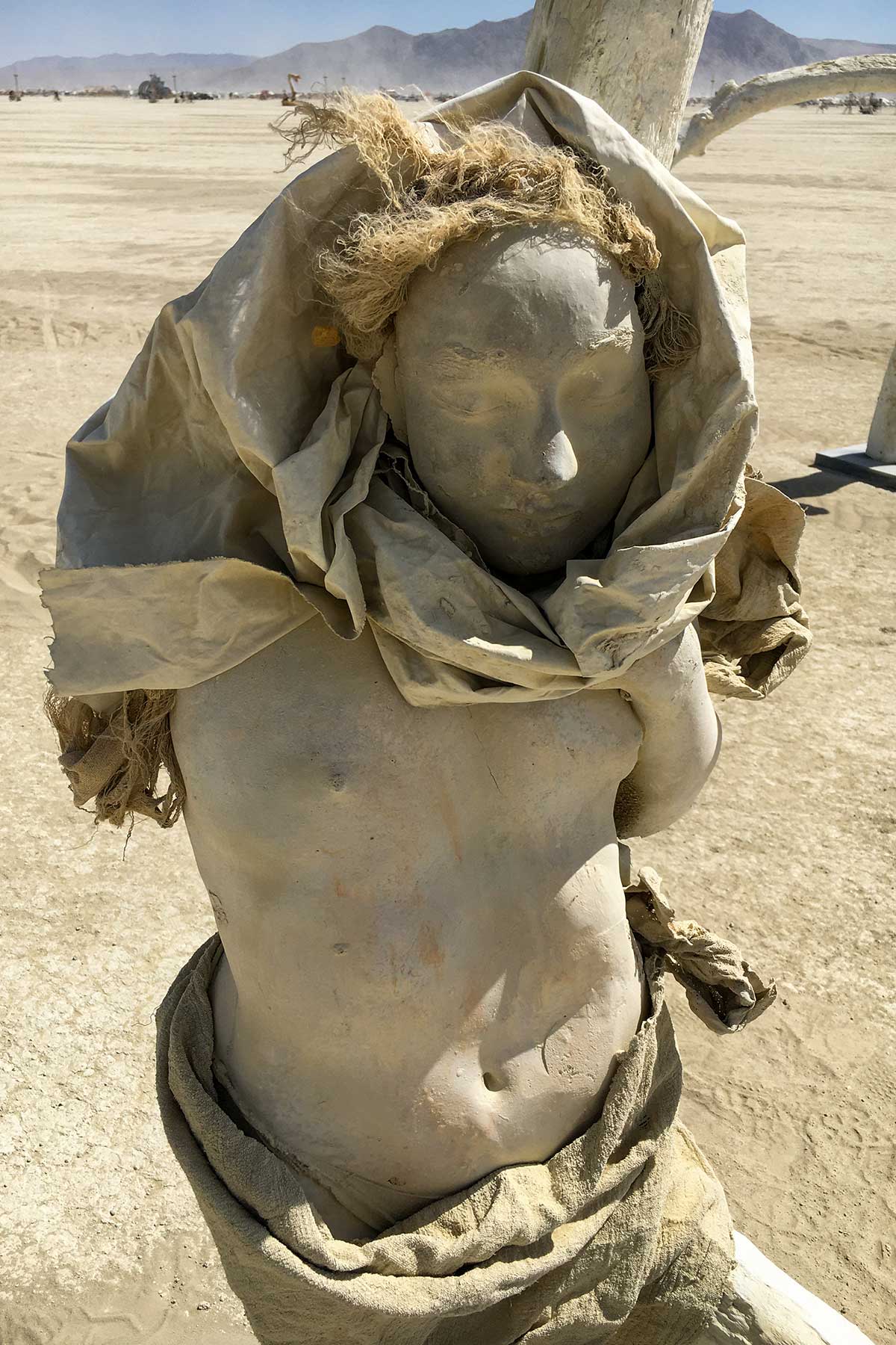 A small hooded cast figure with tufted hair bound to a pale tree, with mountains on the horizon