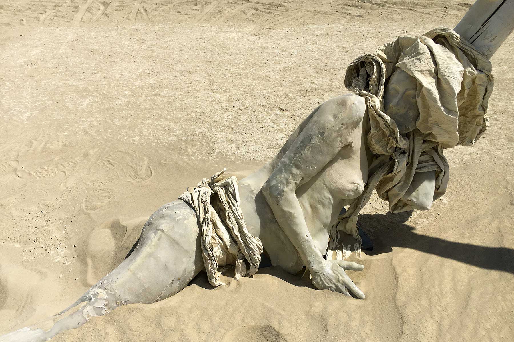 A cast figure half buried in the playa dust, her head wrapped in cloth against a pale tree trunk