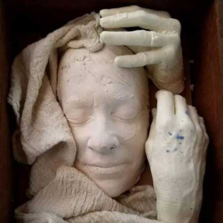 Cast face with closed eyes wrapped in cloth, cradled by two cast hands in a wooden box