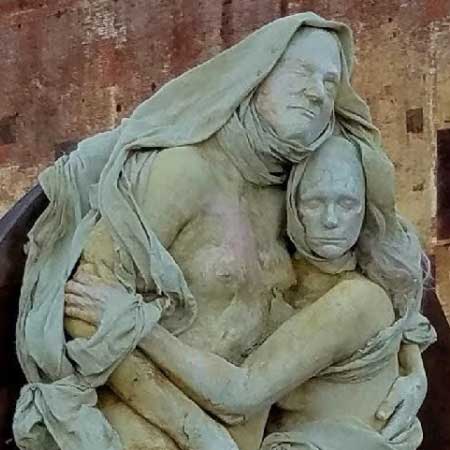 Cast sculpture of a hooded woman embracing a younger figure, both draped in pale green cloth