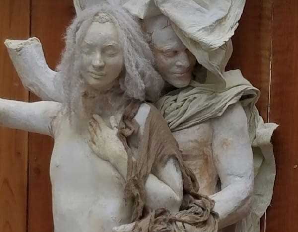 Cast sculpture of a smiling woman with long pale hair, held from behind by a hooded figure